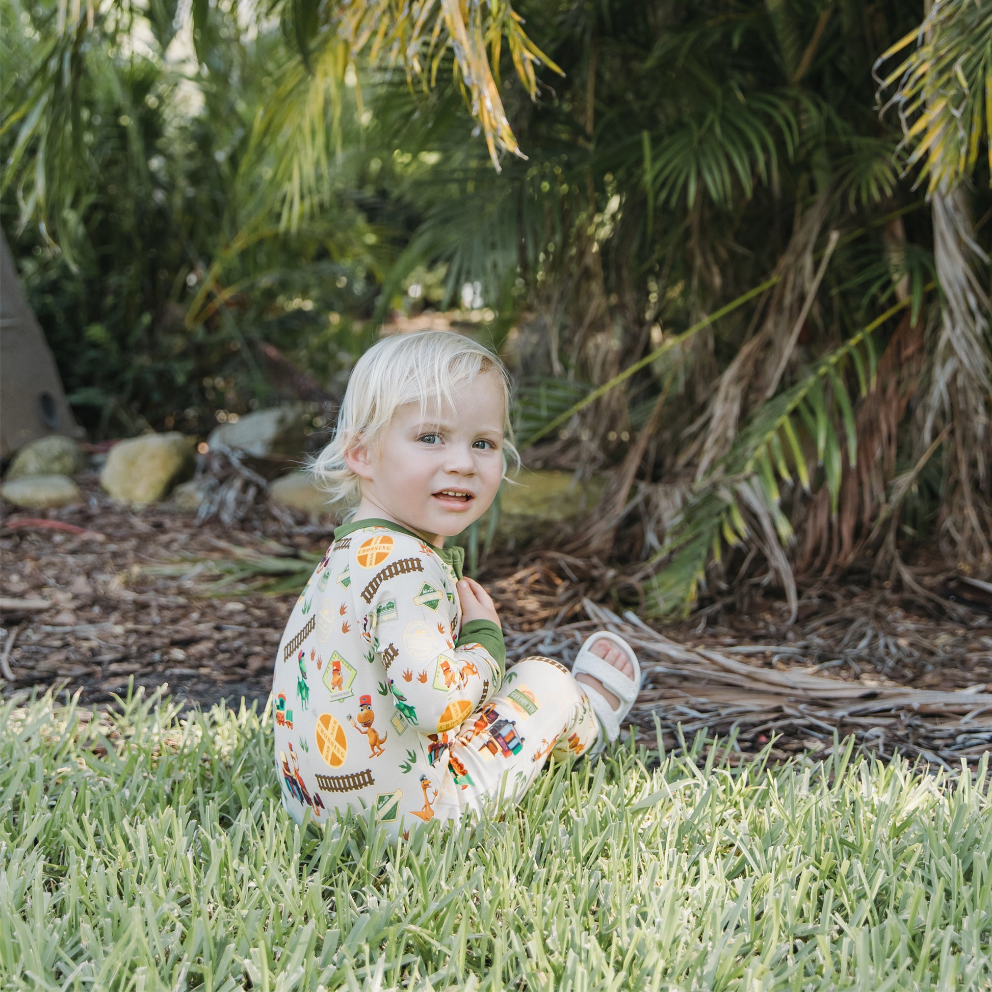 Baby wearing All Aboard the Dinosaur Train™ bamboo pajamas sitting on grass outdoors