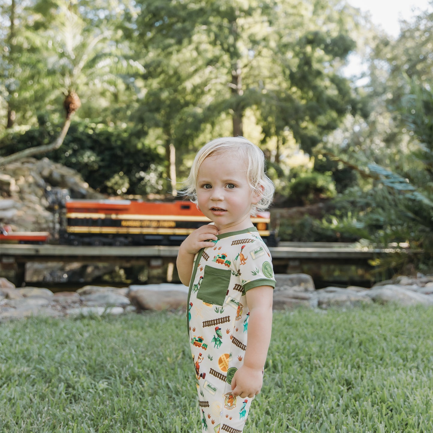 Toddler in All Aboard the Dinosaur Train™ bamboo romper standing near toy train tracks