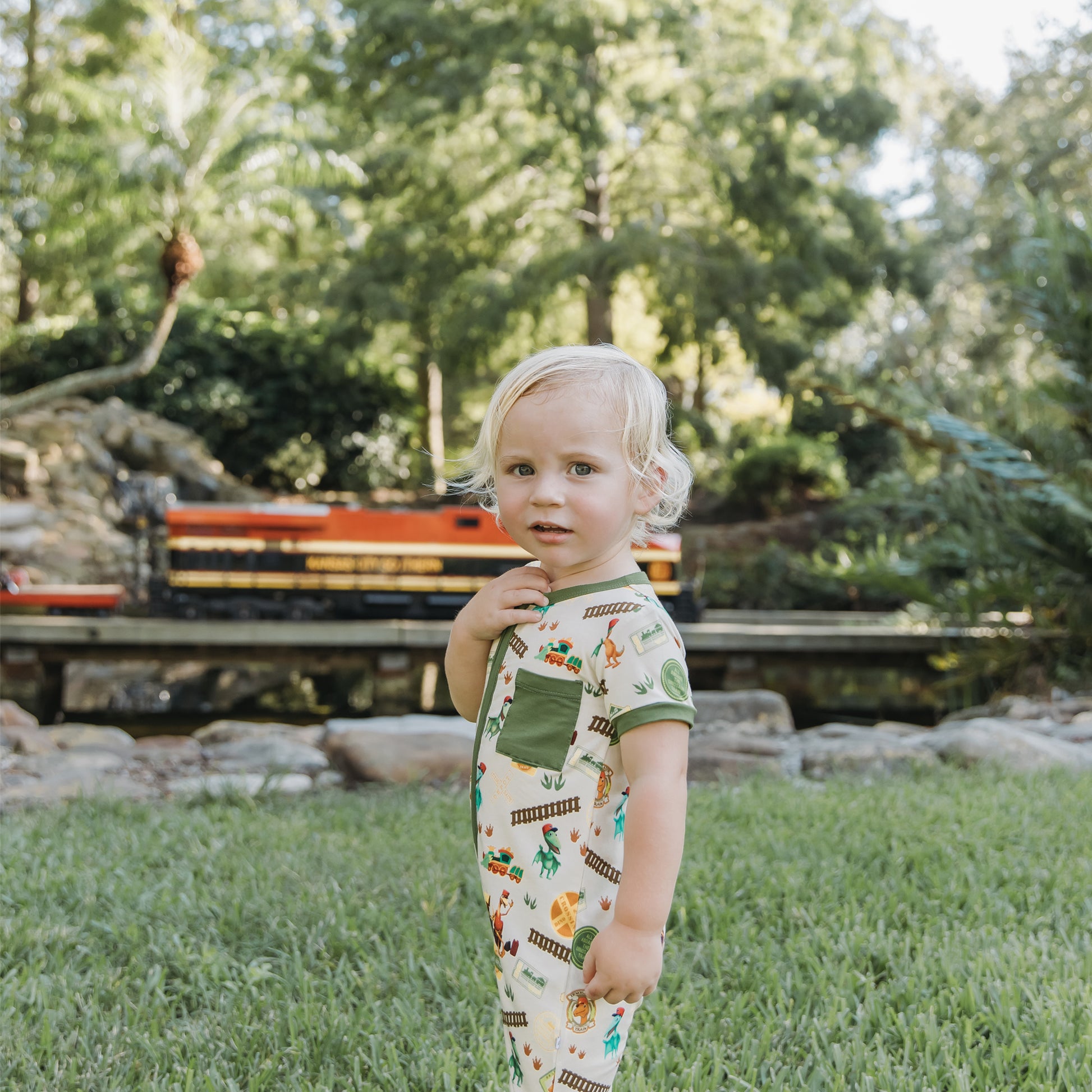 Toddler in All Aboard the Dinosaur Train™ bamboo romper standing near toy train tracks