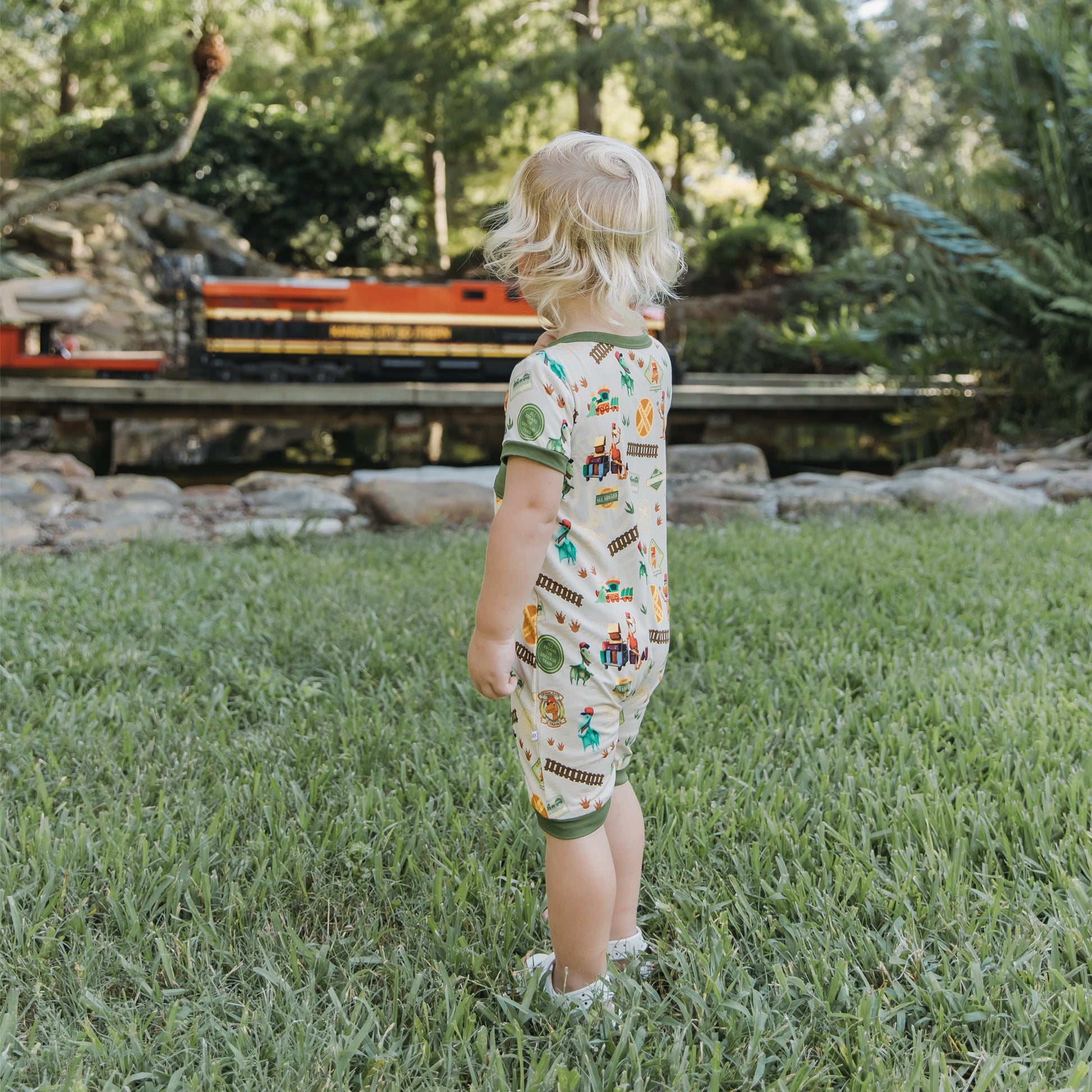 Toddler in All Aboard the Dinosaur Train™ bamboo romper watching a model train outdoors