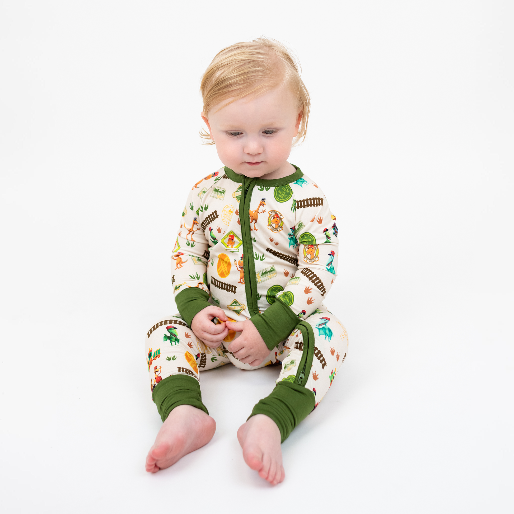 Toddler sitting down with our Emerson and Friends All Aboard the Dino Train bamboo convertible baby pajamas with green trim, double zipper, and colorful orange, blue, and green dinosaurs on train tracks with "Dino Train" print.