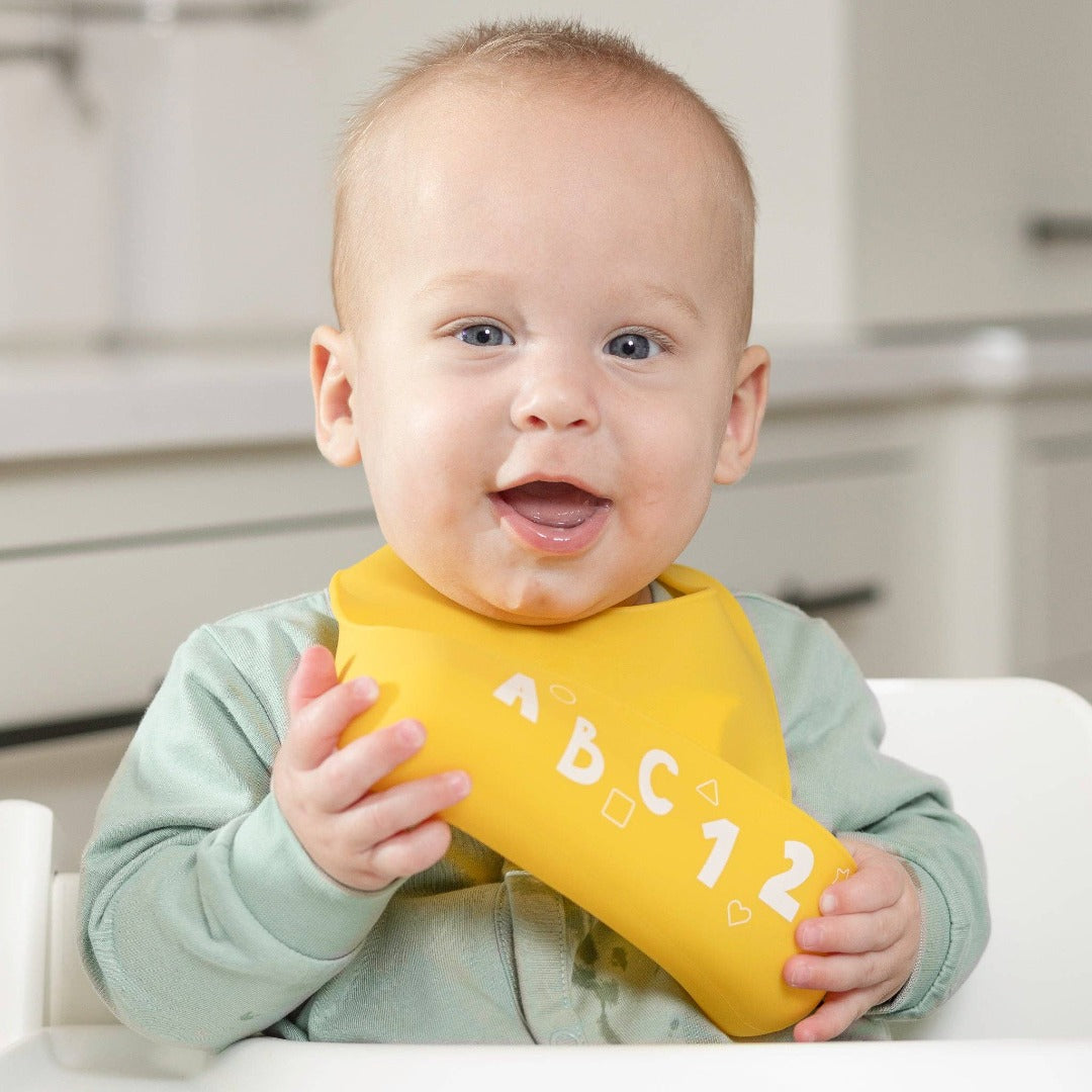 happy baby wearing bright yellow silicone bib with white letters and numbers along the bottom