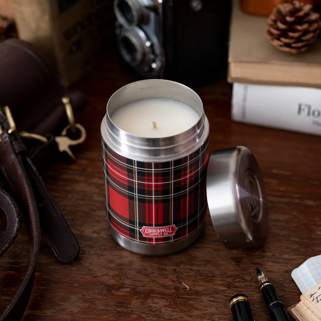 Candle in a plaid container on a wooden surface with a camera and books in the background