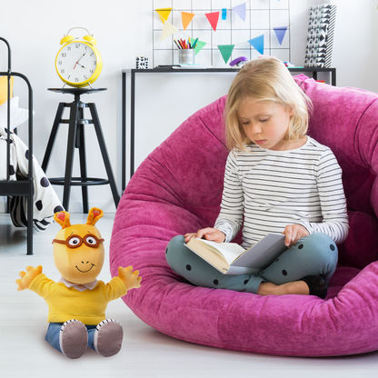 A plush toy of Arthur, a cartoon aardvark, wearing a yellow sweater, blue pants, and red sneakers, waving cheerfully. Besides theres a girl reading in a bean bag.