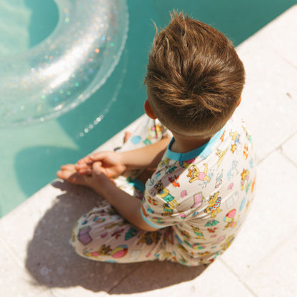 Toddler boy wearing our Arthur's Family Vacation Bamboo Short Sleeve Kids Pajama Pants Set featuring vibrant light blue trim and a playful print of Arthur™ in swimwear surrounded by fun beach props — perfect for family getaways.