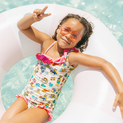 Toddler girl floating in the pool in our Arthur's Family Vacation one piece swimwear featuring vibrant pink trim and the boys swim trunks with a playful print of Arthur™ in swimwear surrounded by fun beach props — perfect for family getaways.