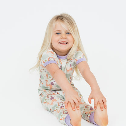 Little girl stretching to reach her toes while sitting in her Magical Unicorn Toddler Bamboo Pajama set. The print includes unicorns, rainbows, clouds, planets, stars, and hearts. The colors are purple, pink, and blu, with hints of green. 