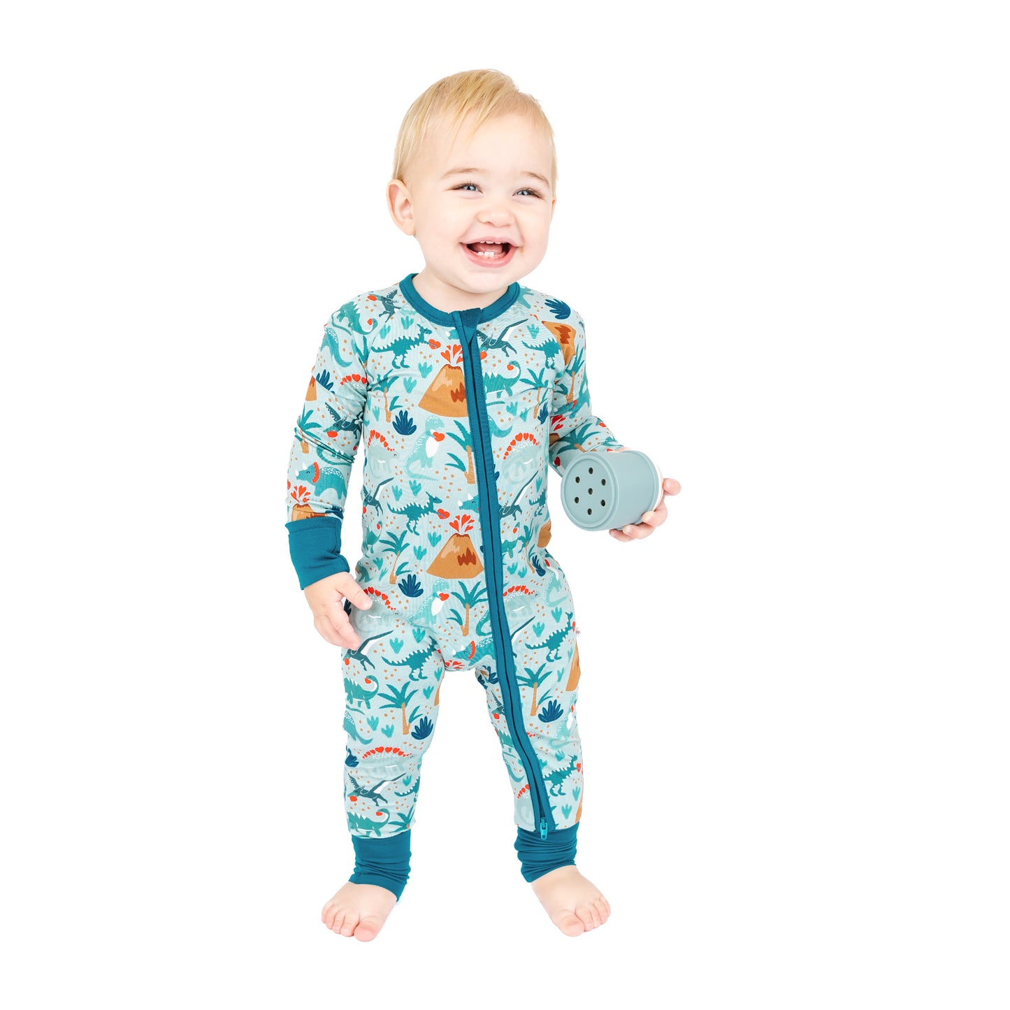 Baby standing in bamboo zip pajama with volcano dinosaur print.