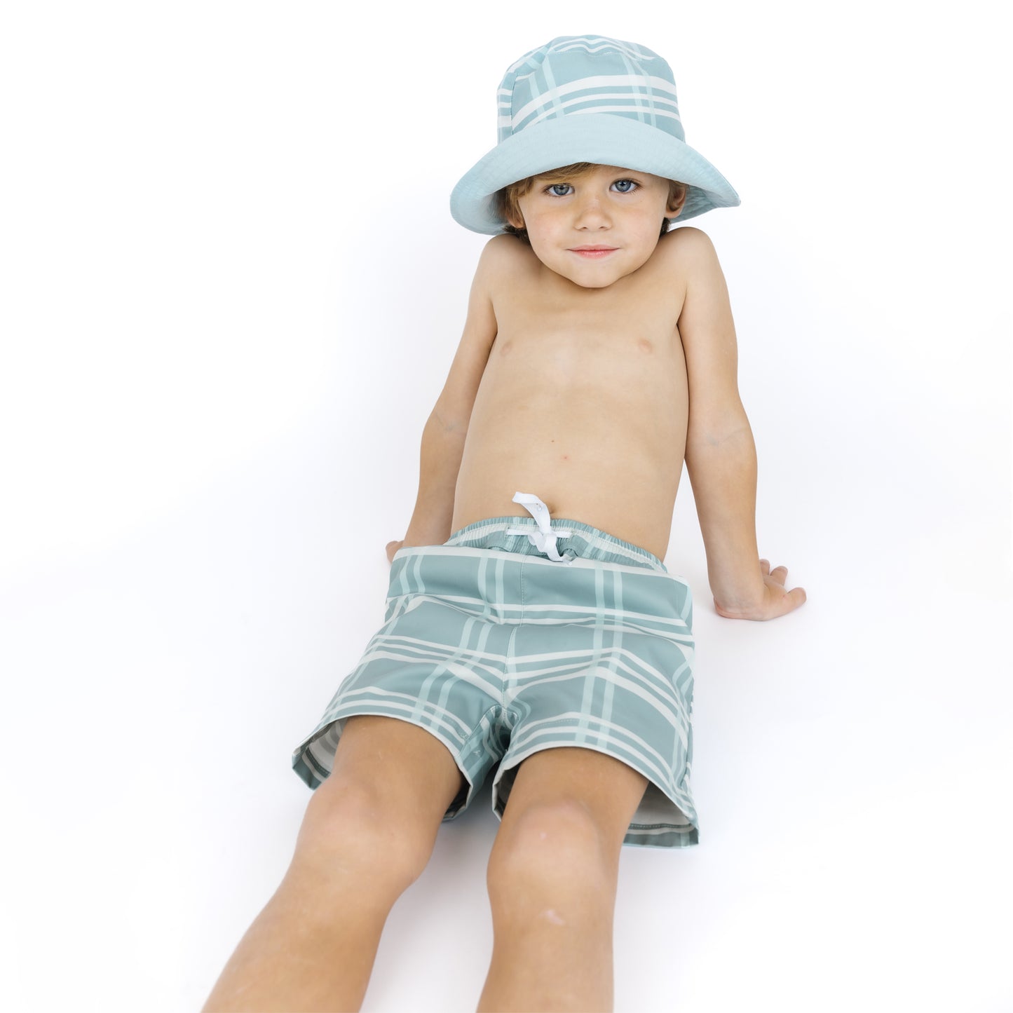 Child wearing a light blue checkered hat and green plaid shorts on a white background