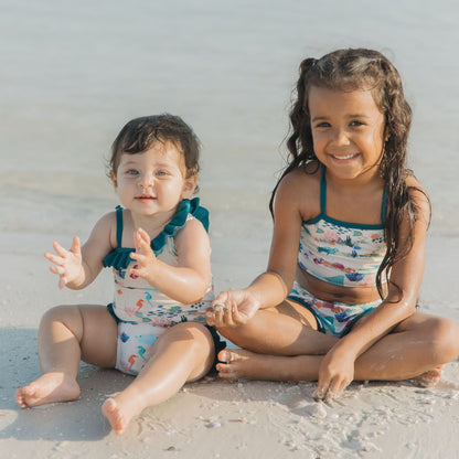 Toddler girl and baby girl wearing a Coral Reef Ruffle Leg One Piece Swimsuit with an ocean-themed print of sharks and sea turtles, designed with soft, stretchy fabric and UPF 50+ sun protection for a fun day at the beach.
