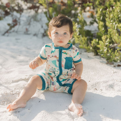 Baby girl sitting at the beach sand wearing Emerson and Friends Coral Reef Bamboo Baby Shortie Rompers, featuring a breathable ocean-themed print with sharks, sea turtles, and coral, designed for summer comfort and easy zip-up wear.
