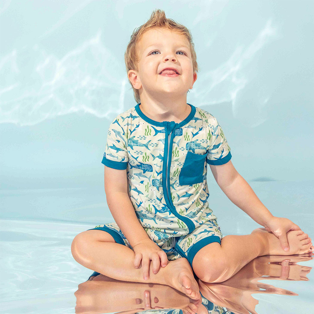 Little boy sitting on the ground with a metallic backdrop wearing our Shark Friends Shortie Romper.