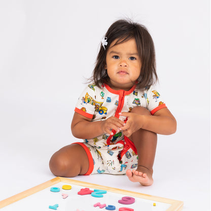 Baby girl sitting playing with letter wearing our soft-buttery bamboo baby shortie romper crafted from buttery-soft bamboo, featuring red trims and playful illustrations of Arthur™ PBS Character, books, and school bus, a beige background.
