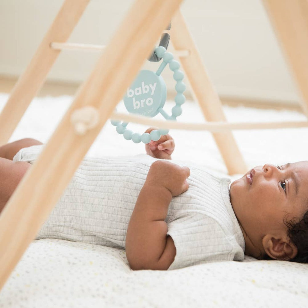 little baby laying on the floor playing with a hanging light blue round teether that reads little bro in white letters