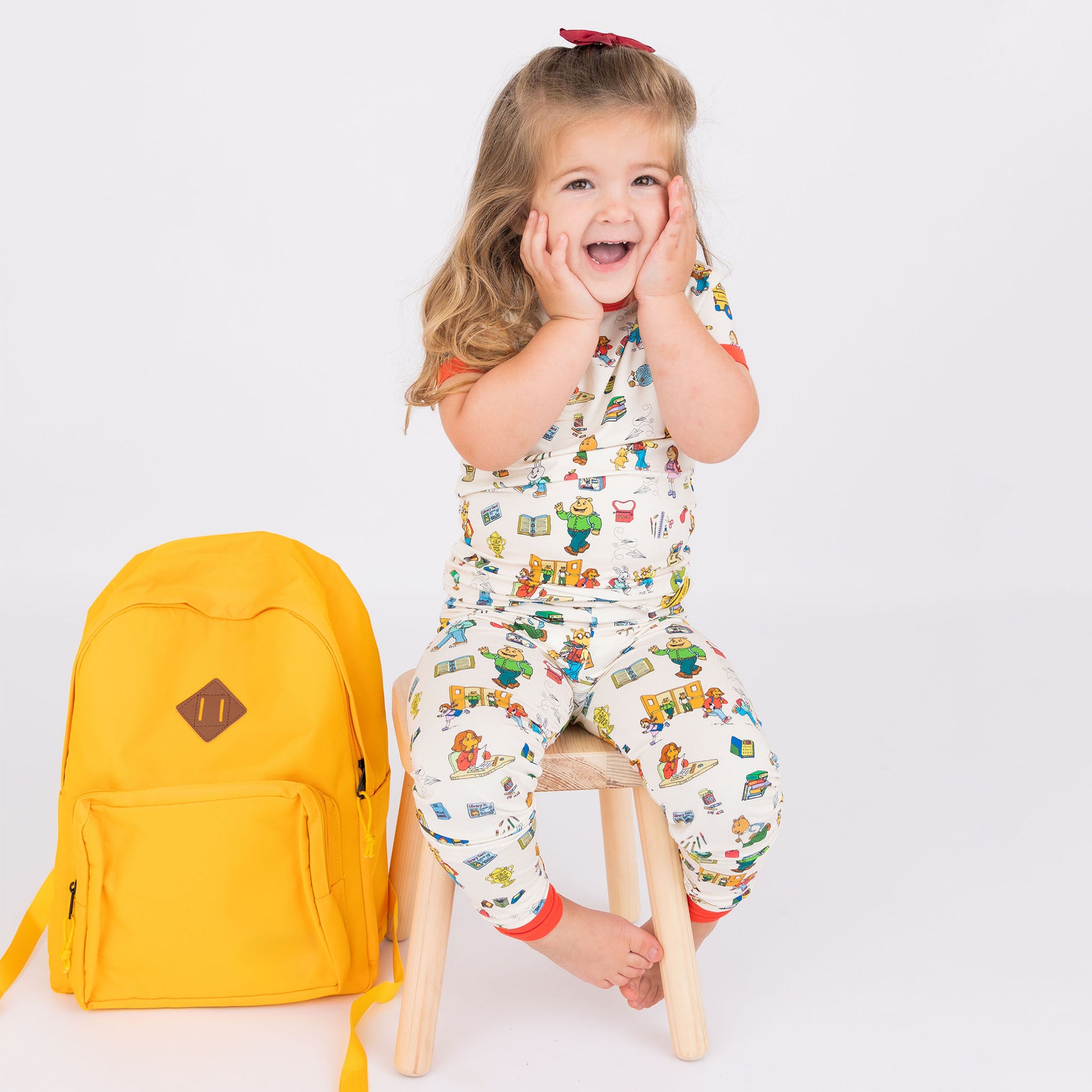 Girl smiling at the camera with a yellow backpack on the side with out short Sleeve Kids Pajama Pants Set from buttery-soft bamboo, featuring red trims and playful illustrations of Arthur™ PBS Character, books, and school bus, a beige background.