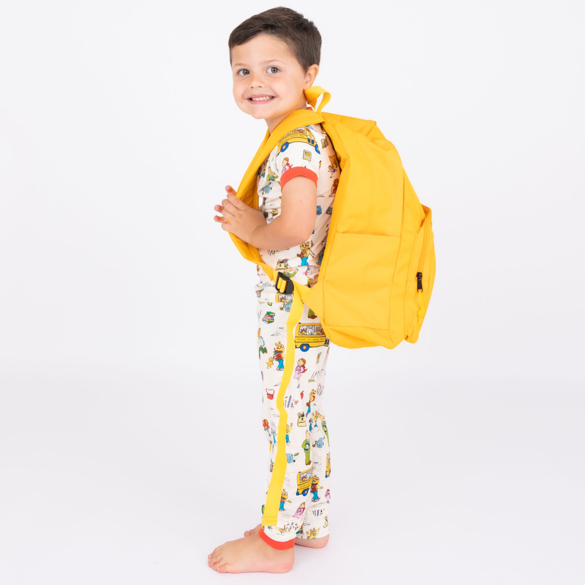 Boy wearing our short Sleeve Kids Pajama Pants Set from buttery-soft bamboo, featuring red trims and playful illustrations of Arthur™ PBS Character, books, and school bus, a beige background. 