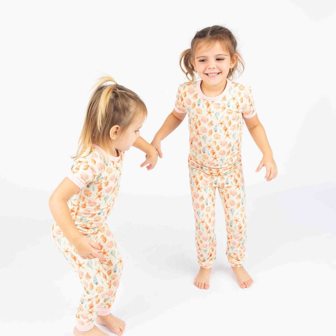 Two toddler girls wearing our 2-piece Sandy Seashell bamboo pajama in a white background. 