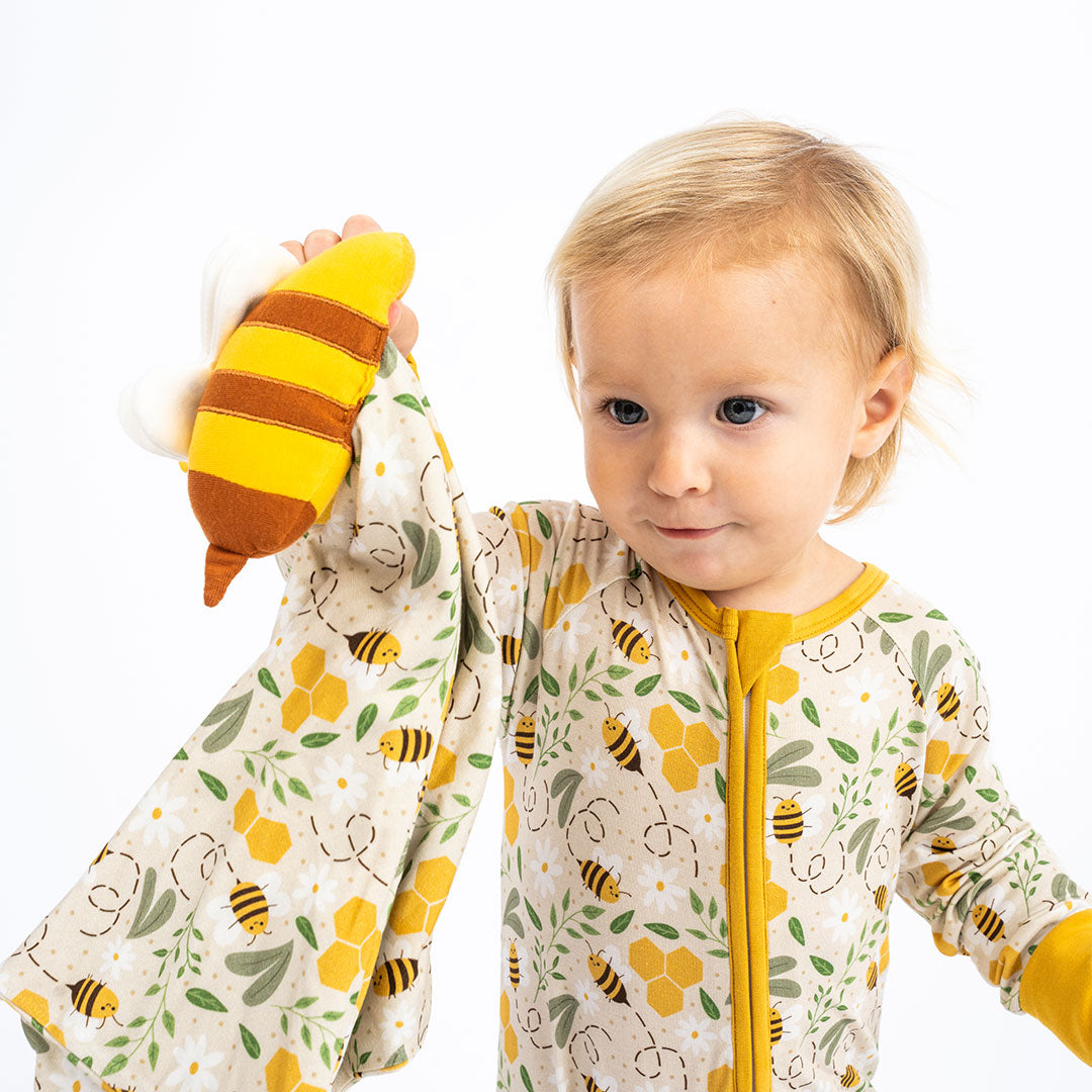 Baby wearing our Happy Honey Bee bamboo pajama with mustard yellow trim. The pajama features a bumblebee, honeycomb, and leaf print, paired with a soft lovey/blankie, on a white background.