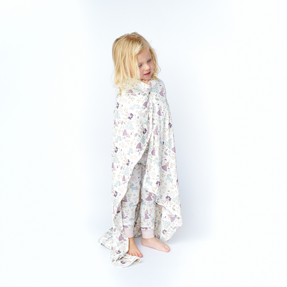 a young girl wraps herself in the "once upon a time" bamboo blanket. the "once upon a time" print is a mix of pinks, purples, whites, blues, and yellows. you can see fairy princess and regular princess, unicorns, swans, and castles. theres also hearts, stars and sparkles scattered around the print. 