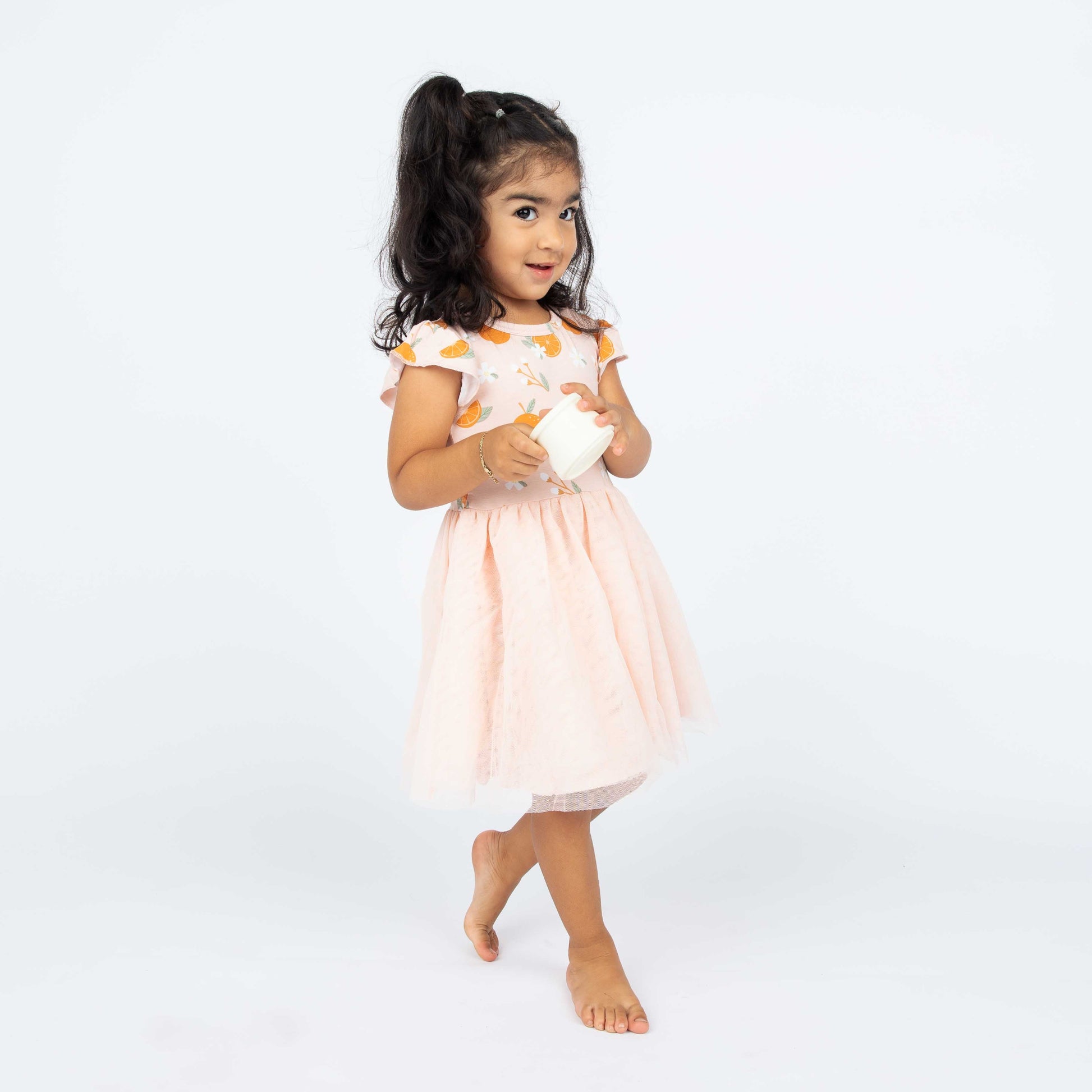 Toddler girl wearing our Freshly Squeezed twirl dress. This dress has the "freshly squeezed" print on the top half and a pink skirt as the bottom half. the "freshly squeezed" print has an assortment of full and half cut oranges scattered around. there is also flower heads and flower stems that intermingle within the print. this is all space out around a pink background space. 