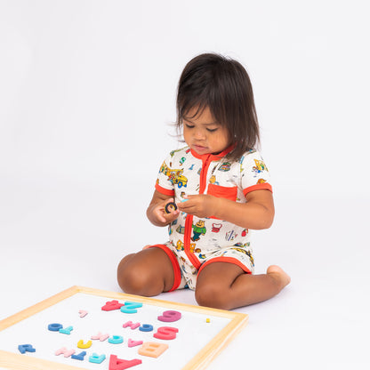 Baby girl sitting playing with letter wearing our soft-buttery bamboo baby shortie romper crafted from buttery-soft bamboo, featuring red trims and playful illustrations of Arthur™ PBS Character, books, and school bus, a beige background.