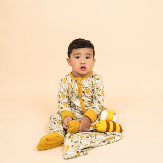 A baby sitting on the floor wearing bee print bamboo pajamas, holding a soft, breathable, hypoallergenic bee-patterned lovey blanket.