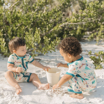 Two babies playing in beach sand wearing Emerson and Friends Coral Reef Bamboo Baby Shortie Rompers, featuring a breathable ocean-themed print with sharks, sea turtles, and coral, designed for summer comfort and easy zip-up wear.