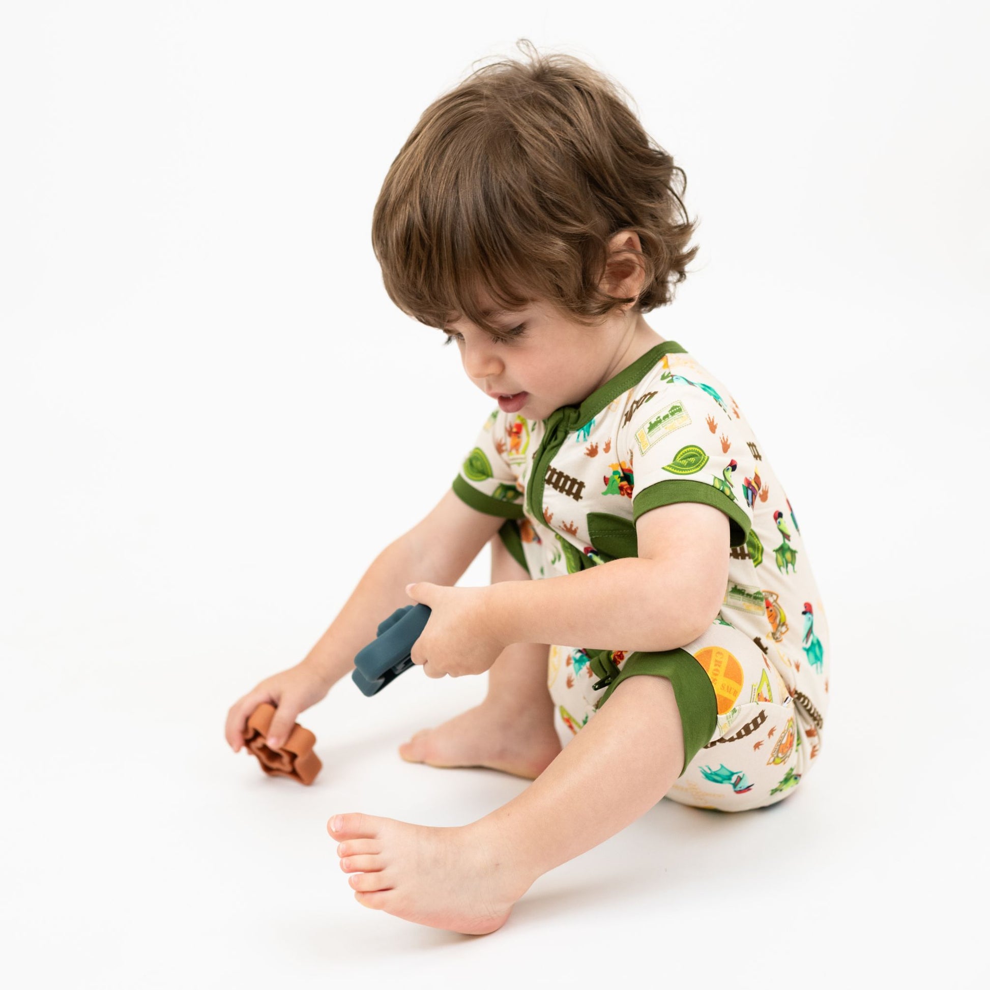 Toddler wearing our Emerson and Friends All Aboard the Dinosaur Train™ Bamboo Baby Shortie Romper featuring with green trim, and colorful orange, blue, and green dinosaurs on train tracks with "Dino Train" print.