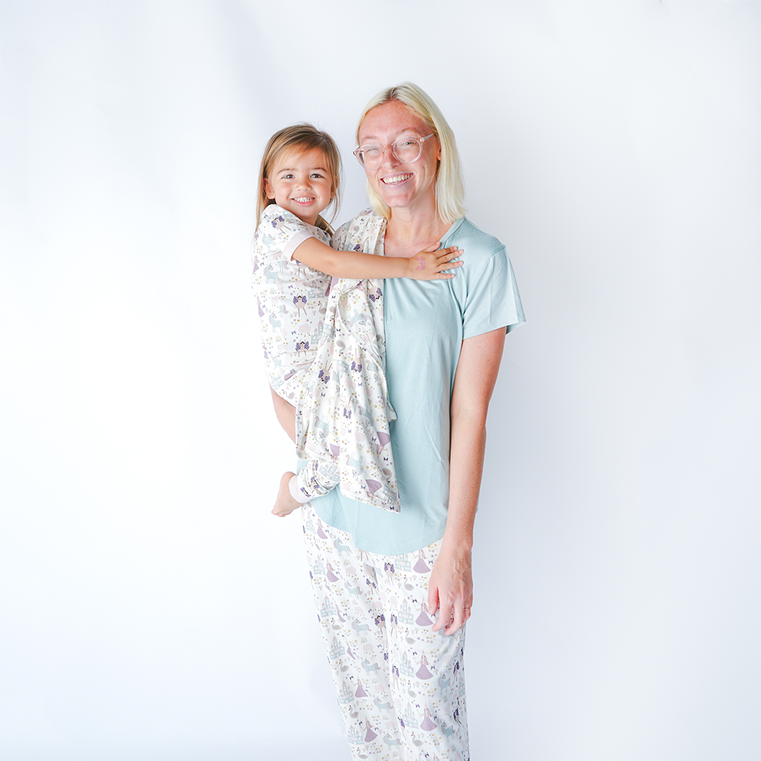 a mom smiles with her daughter on her hip. the "once upon a time" bamboo blanket is draped over her shoulder. they are both wearing "once upon a time" apparel. the mom in the relaxed pants and the daughter in the 2-piece matching pajama set. the "once upon a time" print is a mix of pinks, purples, whites, blues, and yellows. you can see fairy princess and regular princess, unicorns, swans, and castles. theres also hearts, stars and sparkles scattered around the print. 