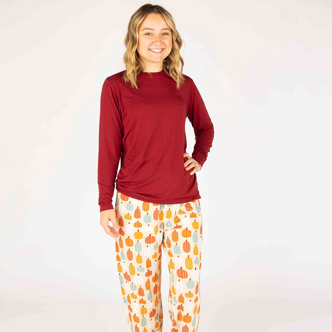 girl smiles in her "pumpkin patch" relaxed pants and dark red long sleeved shirt. the 'pumpkin patch" print is a mix of boho styled and colored pumpkins. you can see white, blue, orange, and red pumpkins in.different shapes and sizes. this is all on a pastel orange background. 