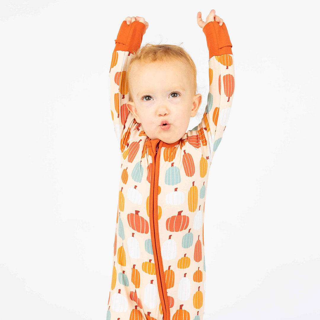 little boy poses with his hands in the air. he wears the "pumpkin patch" convertible. the 'pumpkin patch" print is a mix of boho styled and colored pumpkins. you can see white, blue, orange, and red pumpkins in.different shapes and sizes. this is all on a pastel orange background. 