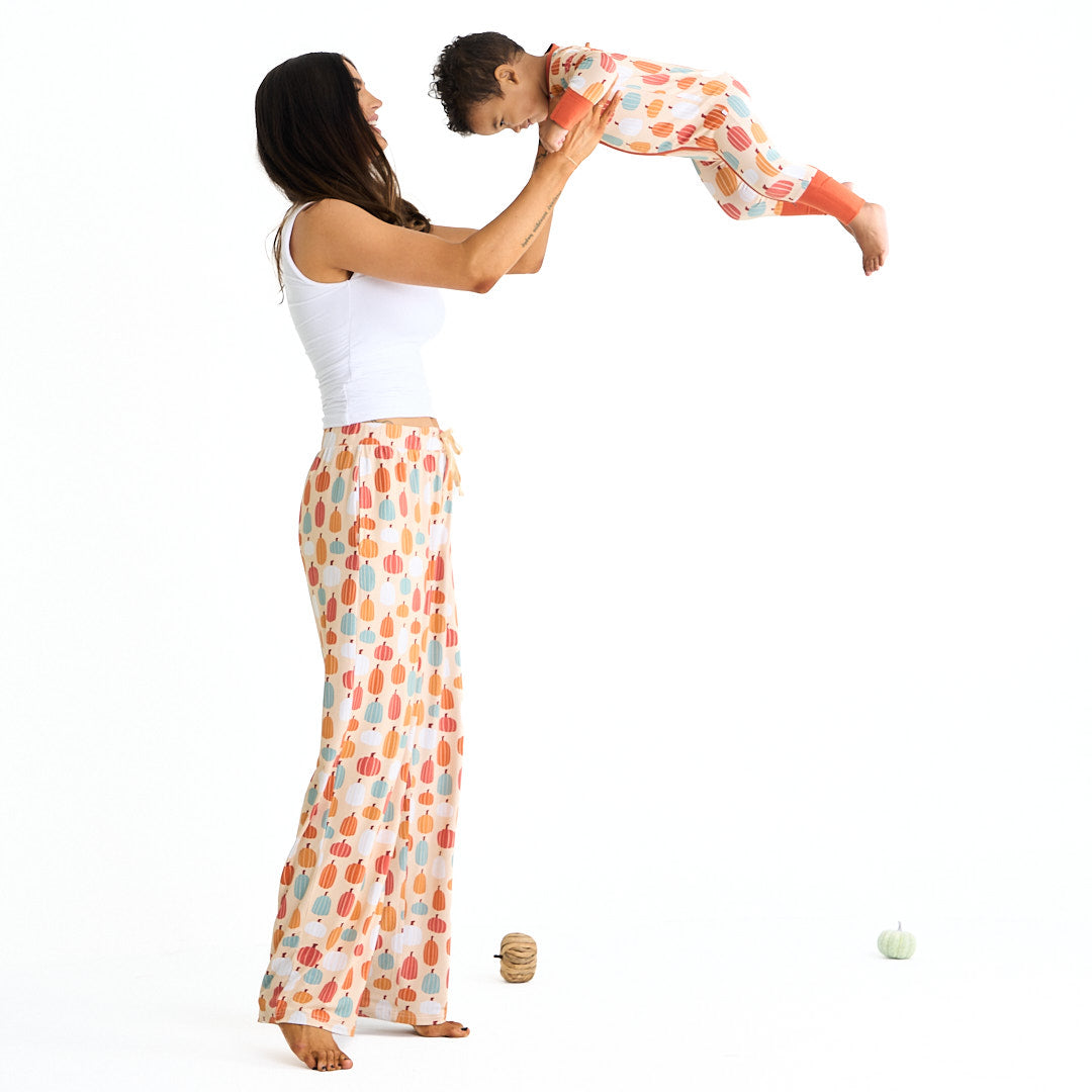 mom holds up and swings her son in the air. the mom wears the "pumpkin patch" relaxed pants. the son wears the "pumpkin patch" convertible. the 'pumpkin patch" print is a mix of boho styled and colored pumpkins. you can see white, blue, orange, and red pumpkins in.different shapes and sizes. this is all on a pastel orange background. 