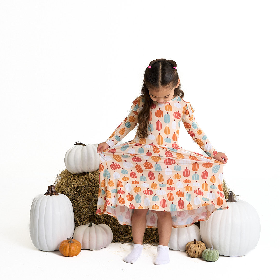 girl stands in front of a pumpkin patch set. she holds the ends of her "pumpkin patch" dress. the 'pumpkin patch" print is a mix of boho styled and colored pumpkins. you can see white, blue, orange, and red pumpkins in.different shapes and sizes. this is all on a pastel orange background. 