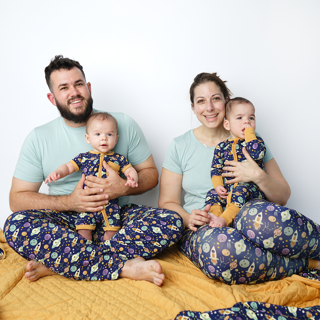 mom and dad sit on the "out of this world" bamboo quilt. they hold their twin babies. they all wear the "out of this world" apparel. dad in the relaxed pants, mom in the joggers, twin 1 in the shortie and twin 2 in the convertible. the "out of this world" print is an outer space theme. you can find spaceships, alien ships, planets, constellations, planets, stars, and moons. 