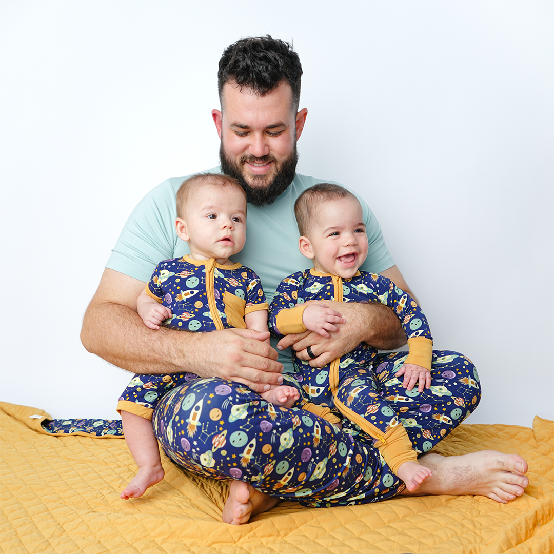 a dad sits on the floor with his twin babies. they are all in "out of this world" attire. 1 twin in the shortie, the other in the convertible. he wears the relaxed pants. the "out of this world" print is an outer space theme. you can find spaceships, alien ships, planets, constellations, planets, stars, and moons. 