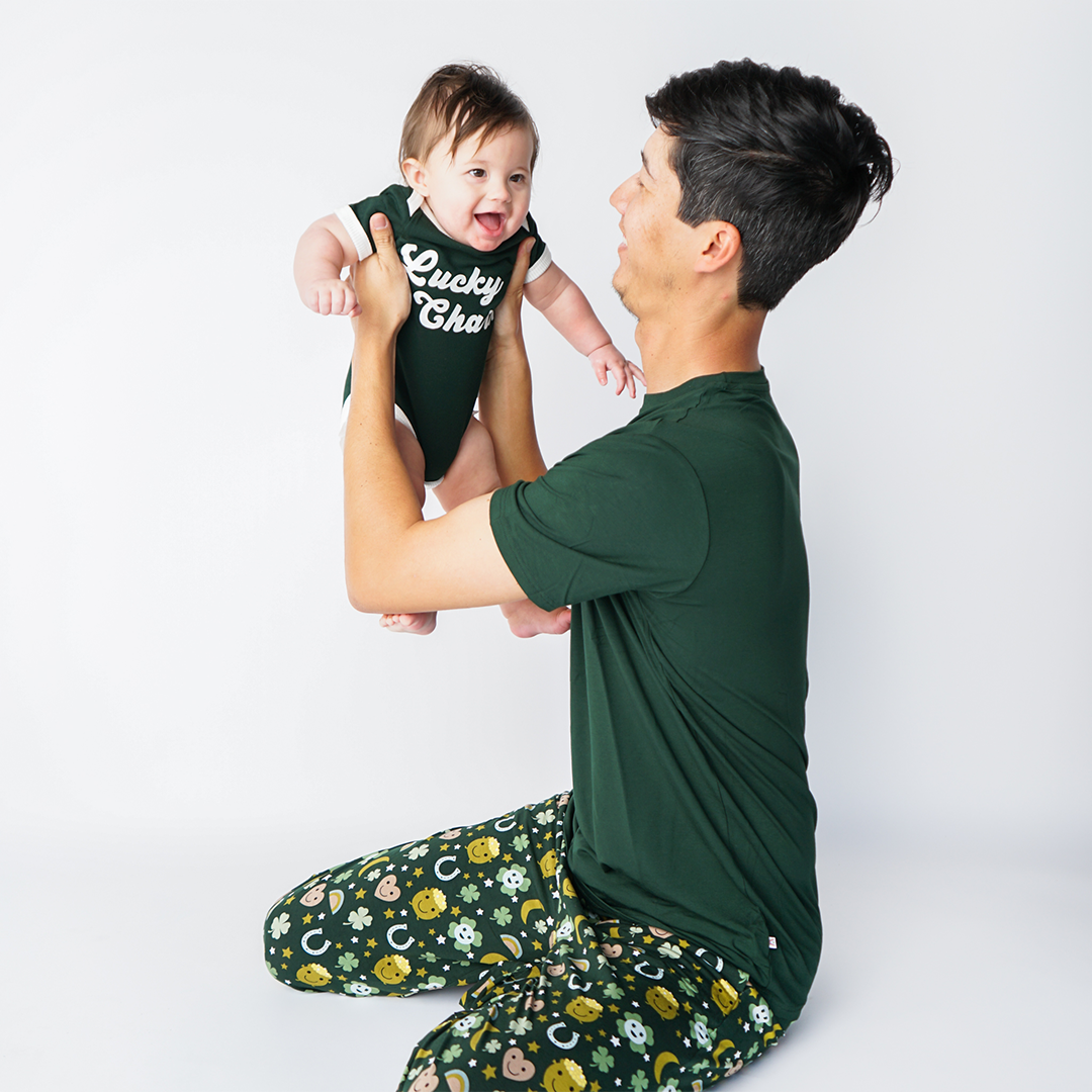 father sits on the floor and holds his baby in the air. the baby wears the "lucky charm" green bamboo terry onesie. 