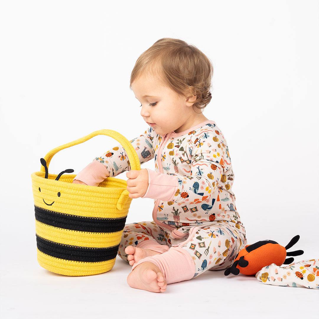 A toddler in a cozy bamboo convertible pajama set reaches into a yellow and black bee-themed rope basket, with a soft ladybug plush toy nearby.