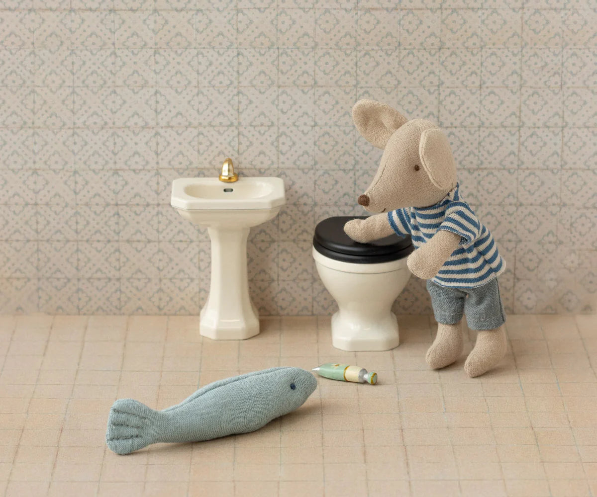 Stuffed toy mouse on a toilet with a fish and bottle on the floor, in front of a sink.