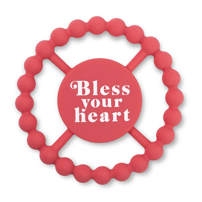pink round silicone teether that says bless your heart on the front in white letters