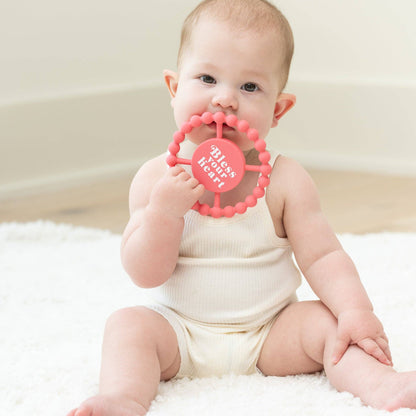 little baby with pink round silicone teether that says bless your heart on the front in white letters