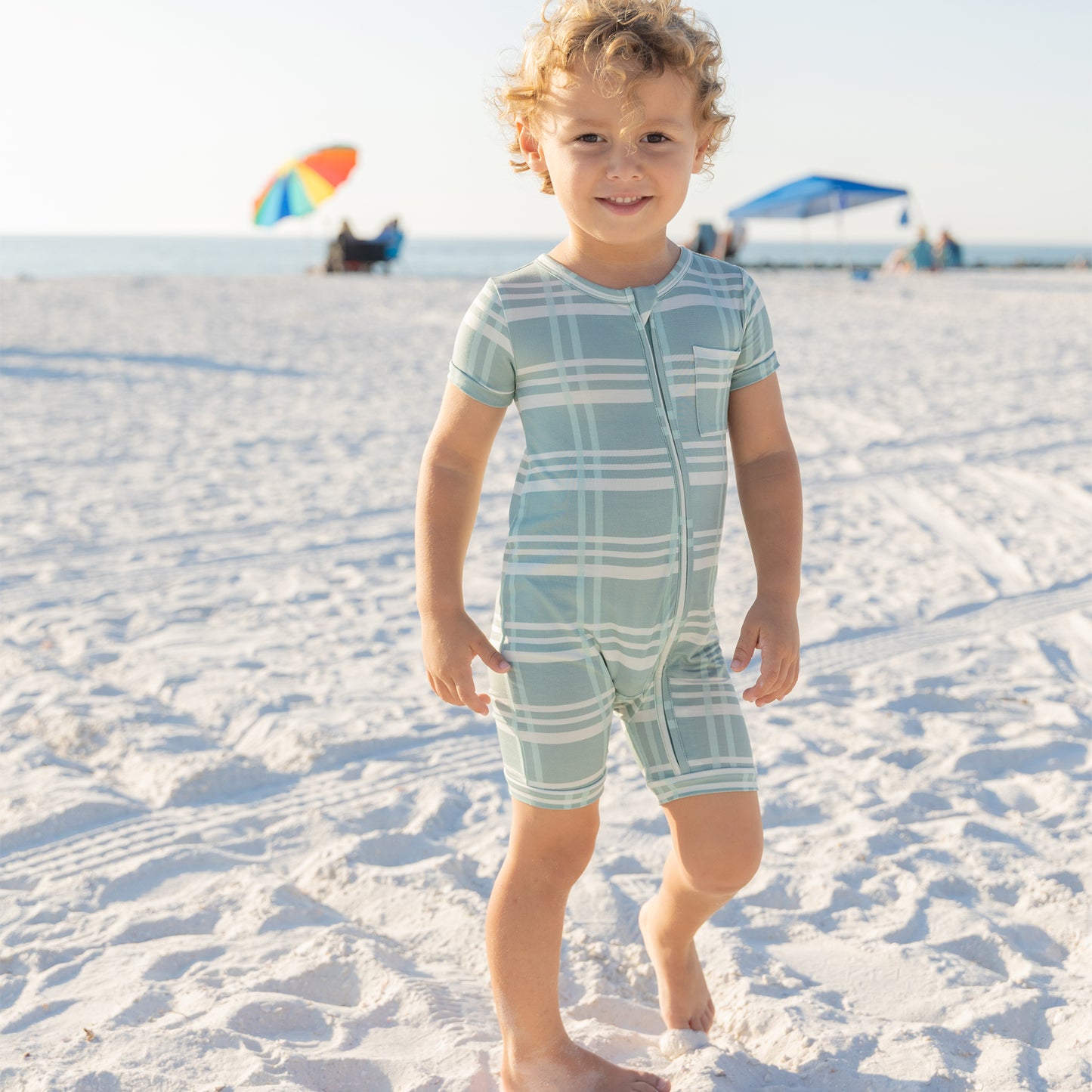 Child in a blue and white checkered romper standing on a sandy beach with umbrellas in the background.