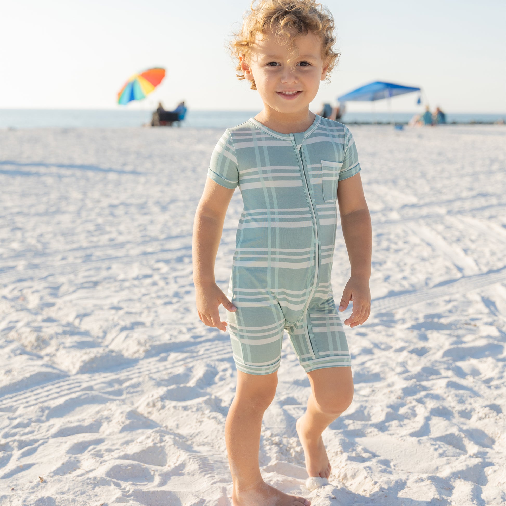 Child in a blue and white checkered romper standing on a sandy beach with umbrellas in the background.