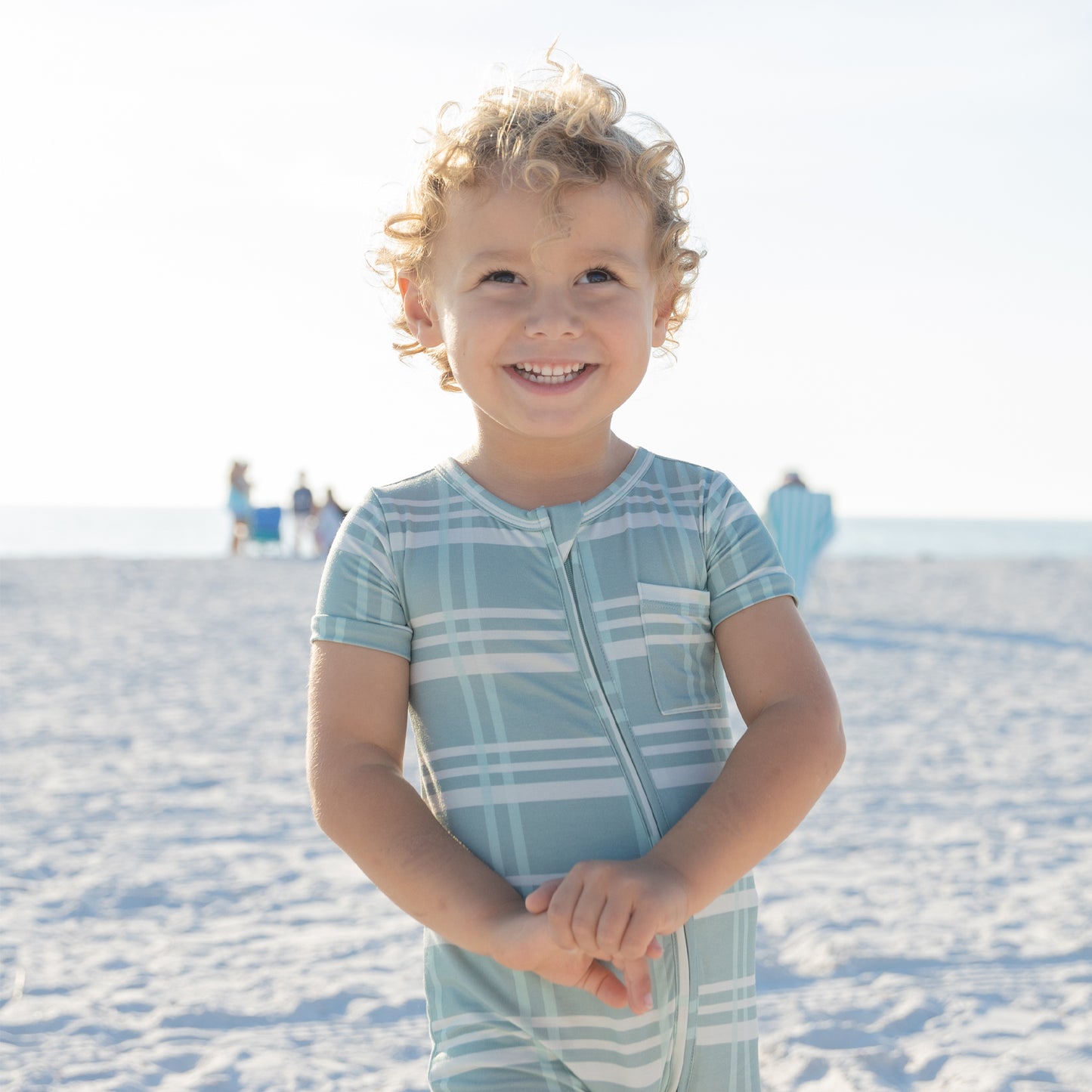 Child wearing a light blue and white checkered outfit on a sandy beach.