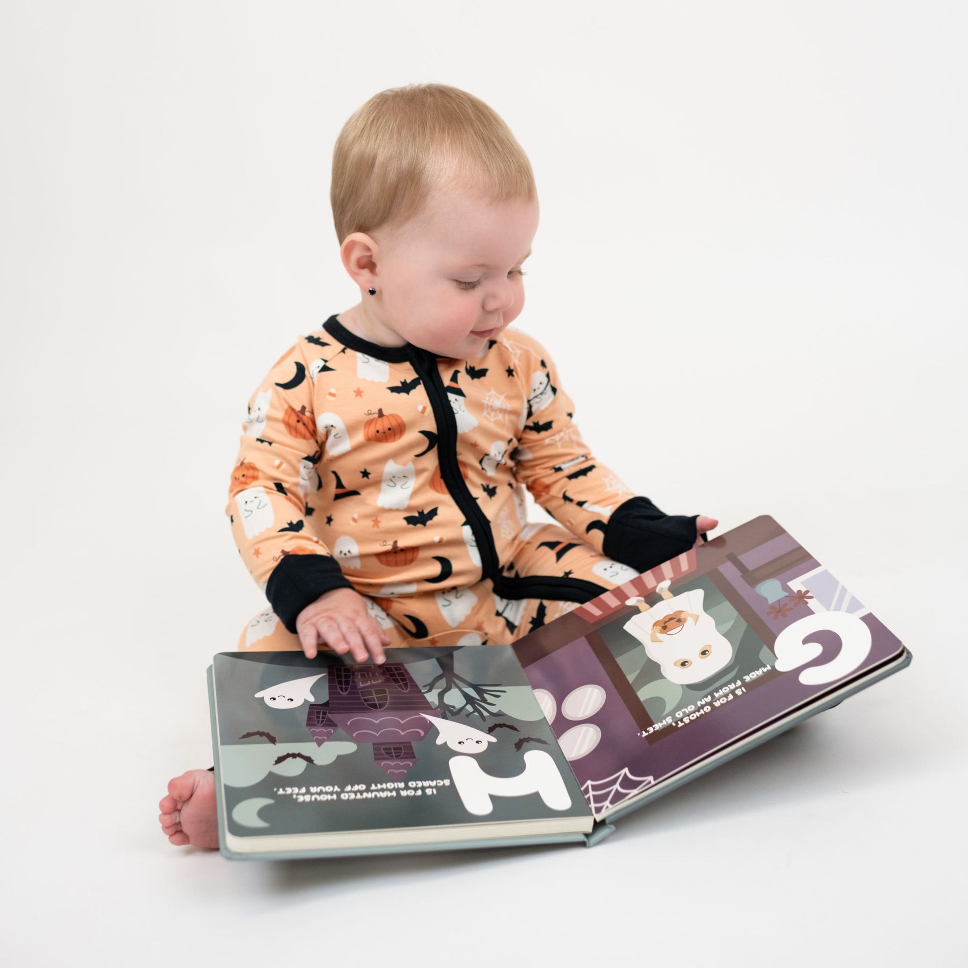 Baby wearing Boo-Tastic Halloween bamboo convertible double zipper pajamas with ghost and pumpkin print, sitting and reading a Halloween board book.