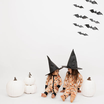 Kids wearing Boo-Tastic Halloween bamboo pajamas sitting with witch hats, white pumpkins, and bat wall backdrop.