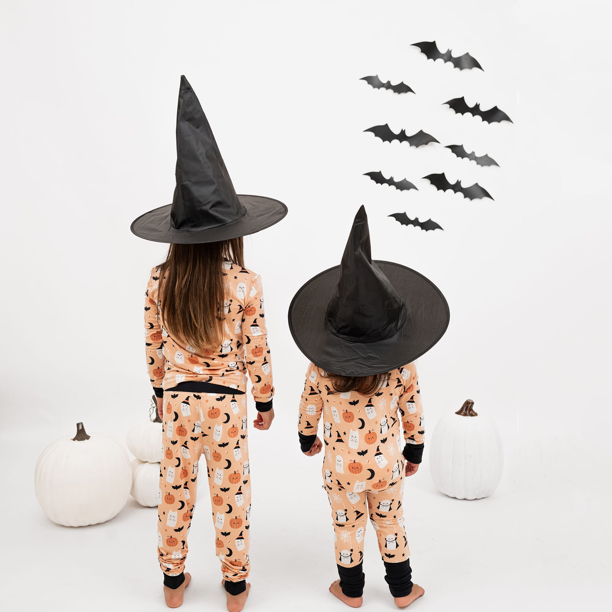 Two kids wearing Boo-Tastic Halloween bamboo pajamas, facing back with witch hats, white pumpkins, and bat wall decor.