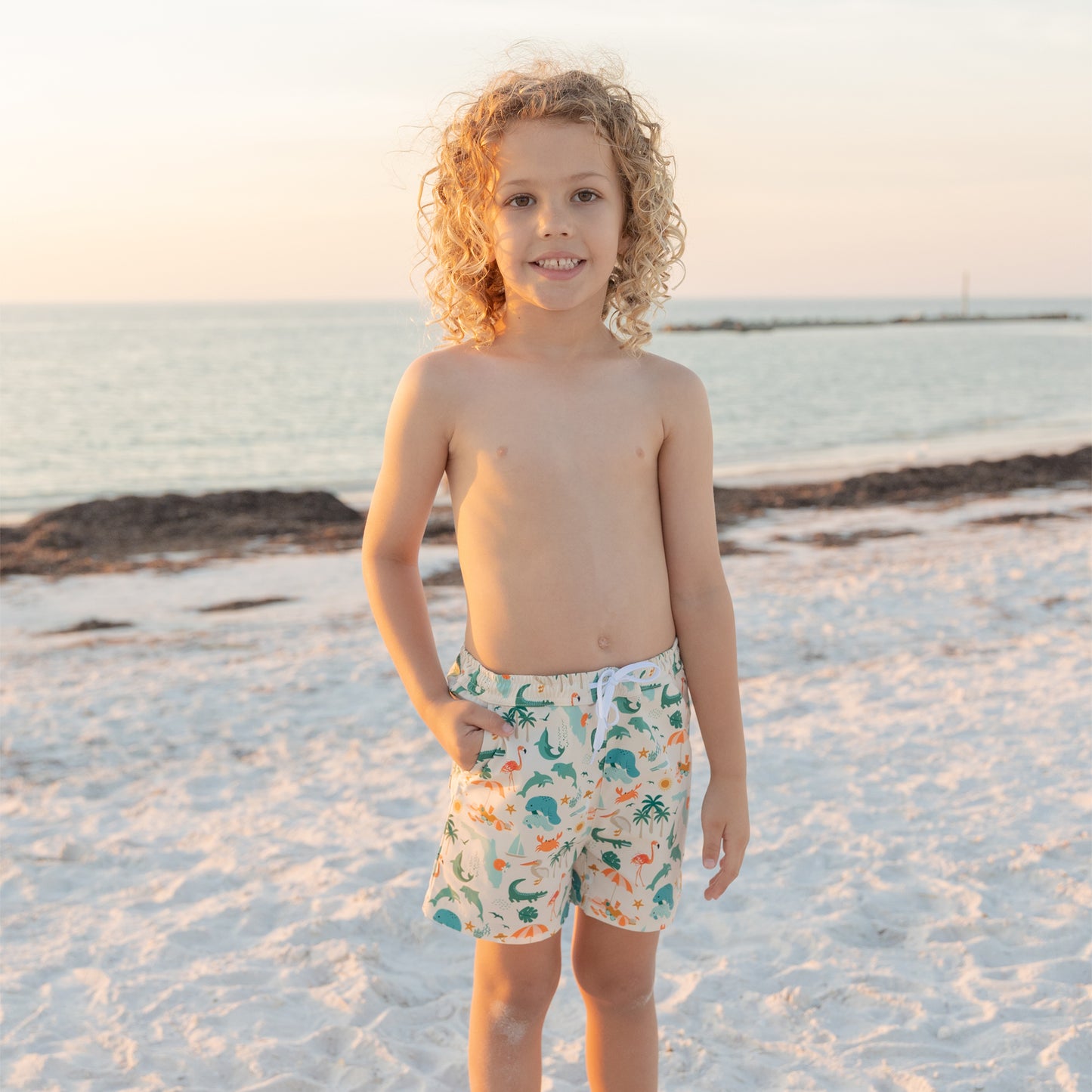 A young boy standing on a beach at sunset wearing Explore Florida swim trunks, showing the relaxed and comfortable fit.
