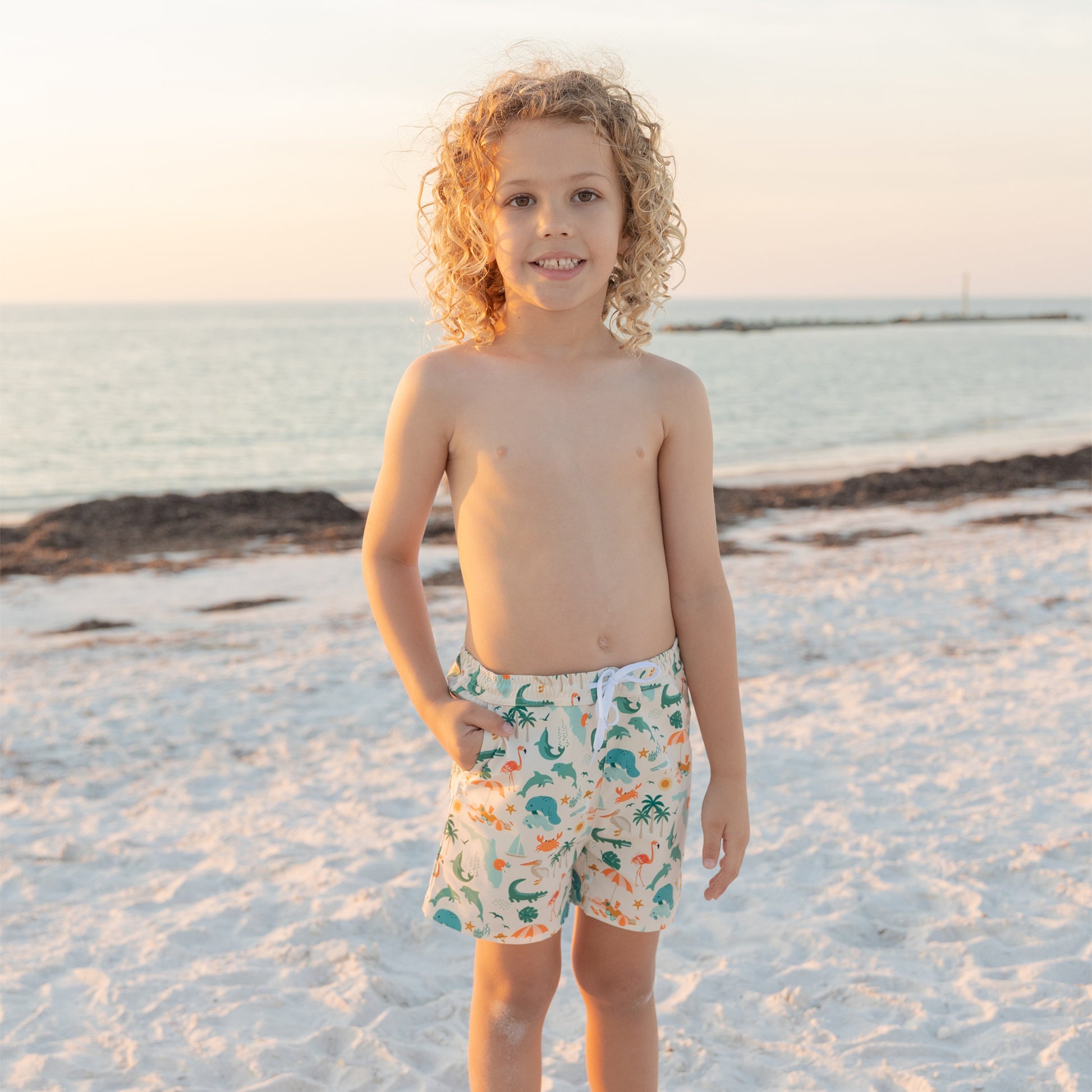 A young boy standing on a beach at sunset wearing Explore Florida swim trunks, showing the relaxed and comfortable fit.