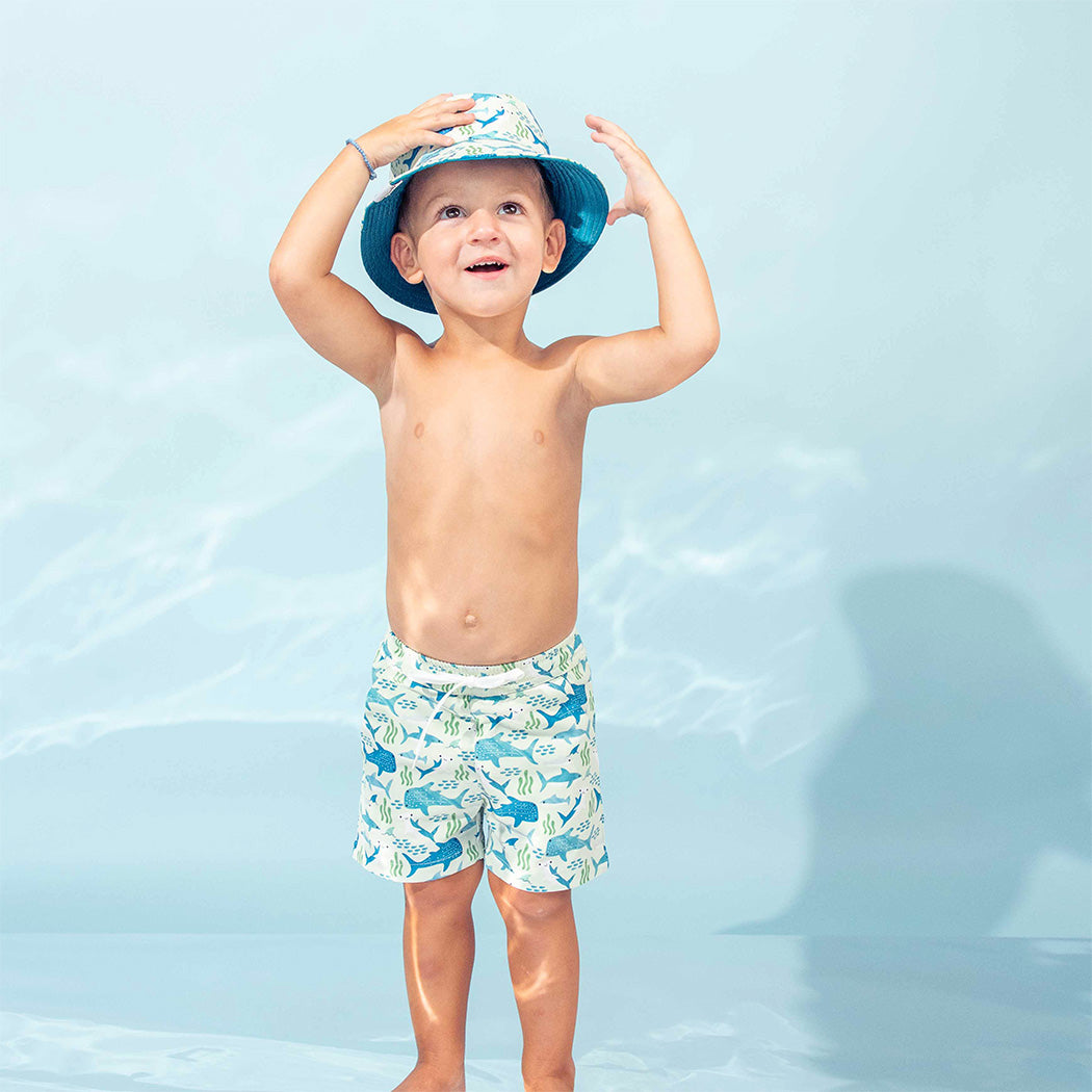 Little boy wearing our Shark Friends Boys Swim Trunks and Bucket Hat in front of the metallic under the sea type backdrop.