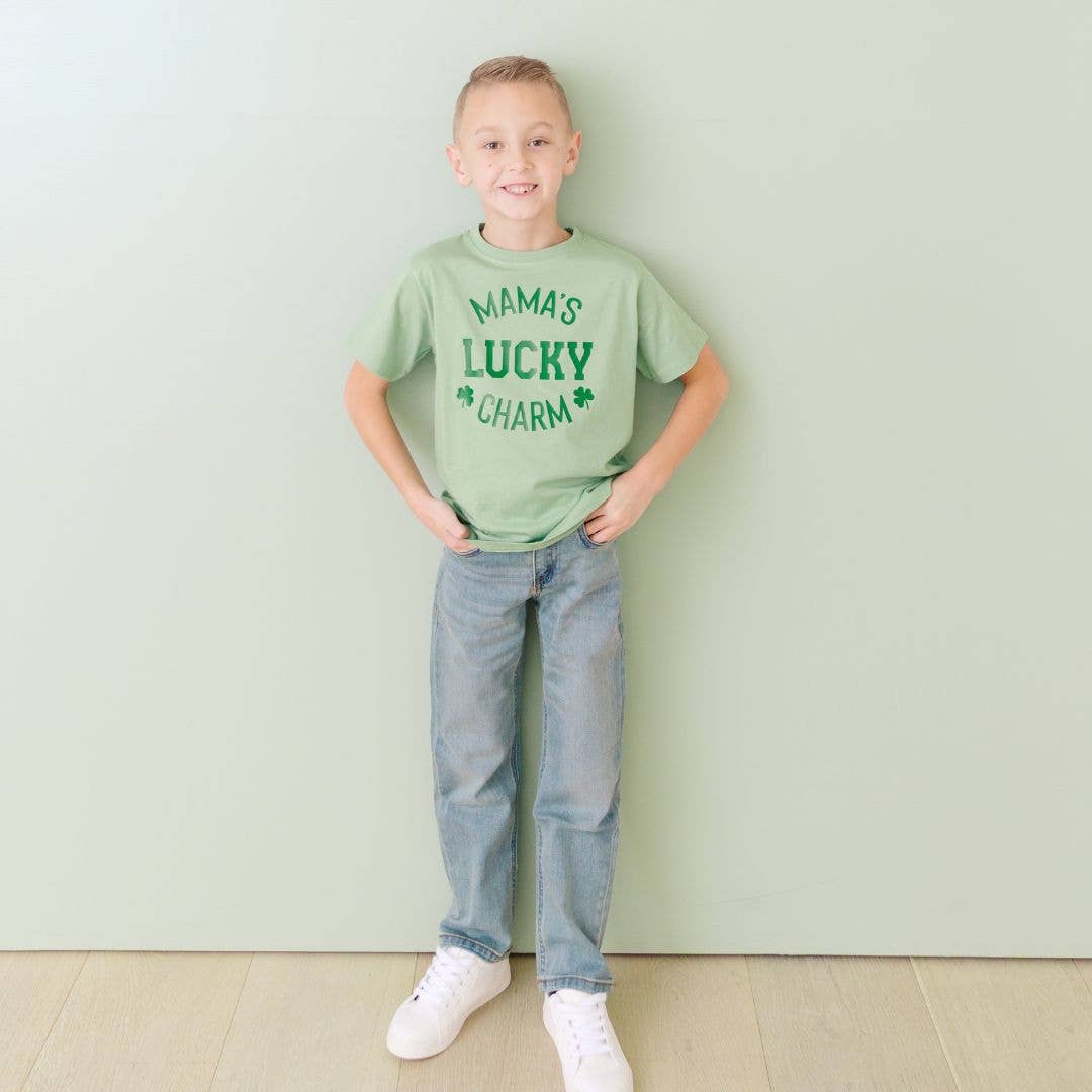 Child wearing a green t-shirt with 'Mama's Lucky Charm' text, standing against a light green wall.